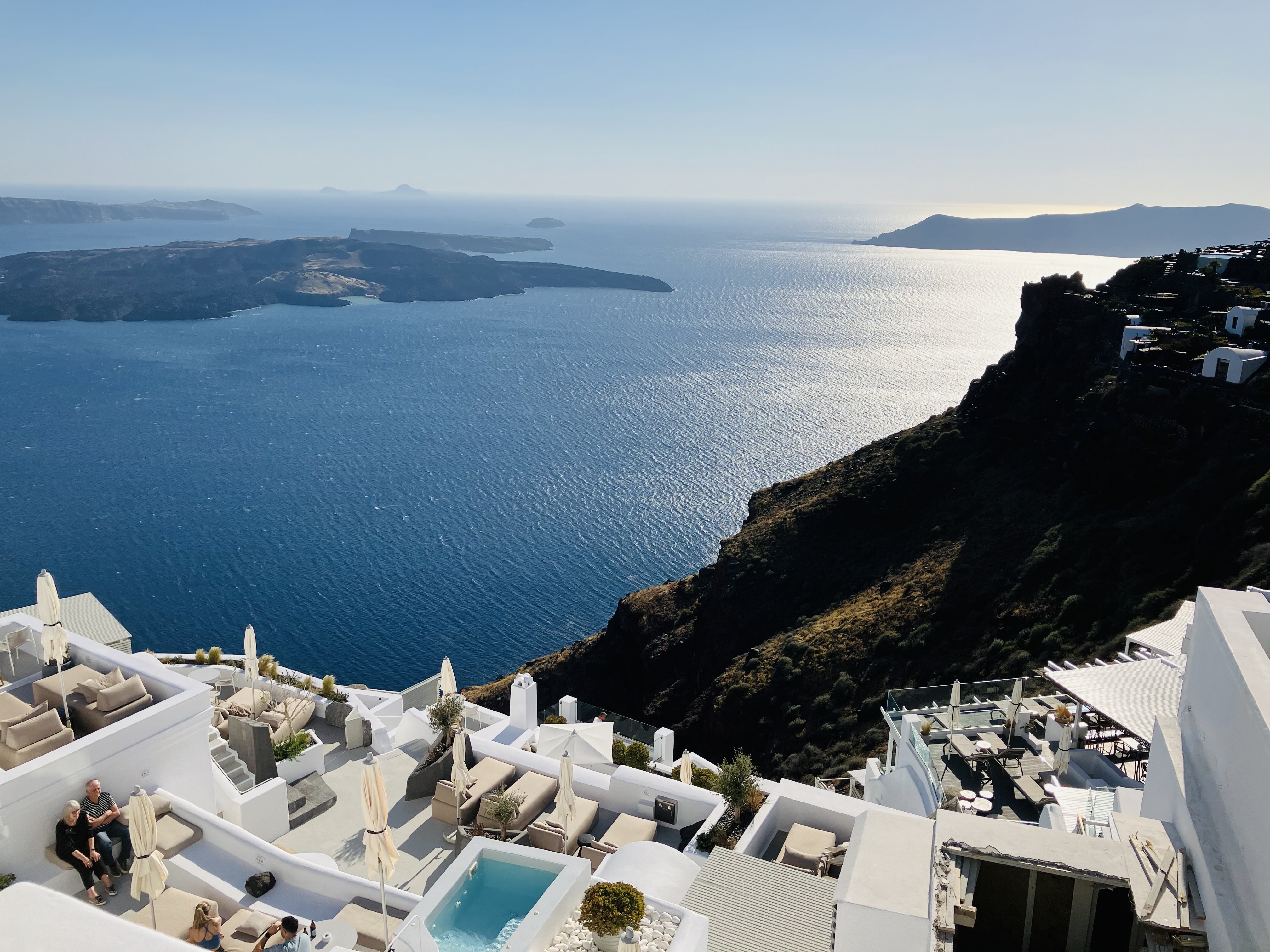 Überdachte Terrasse mit zwei Stühlen und Tisch vor Infinity Pool mit Ausblick auf das Meer, welches von einem Nadelbaum verdeckt wird